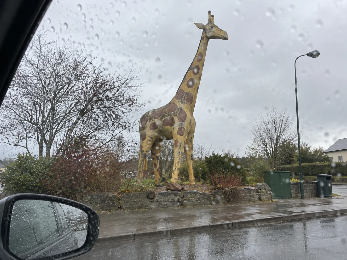 A weathered roadside giraffe sculpture viewed through a rain-speckled car window.