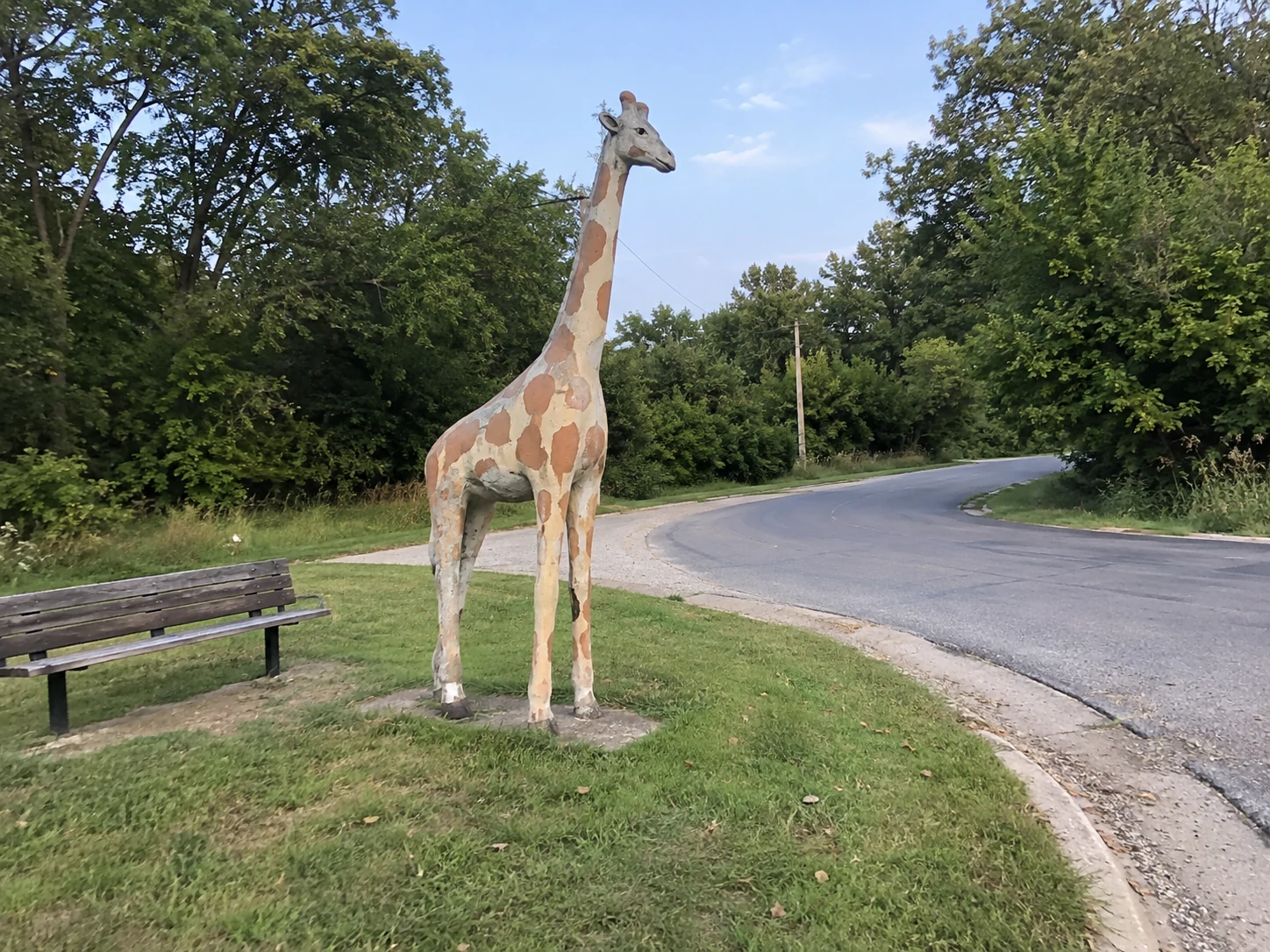 A modest giraffe statue and bench beside an empty road corner.