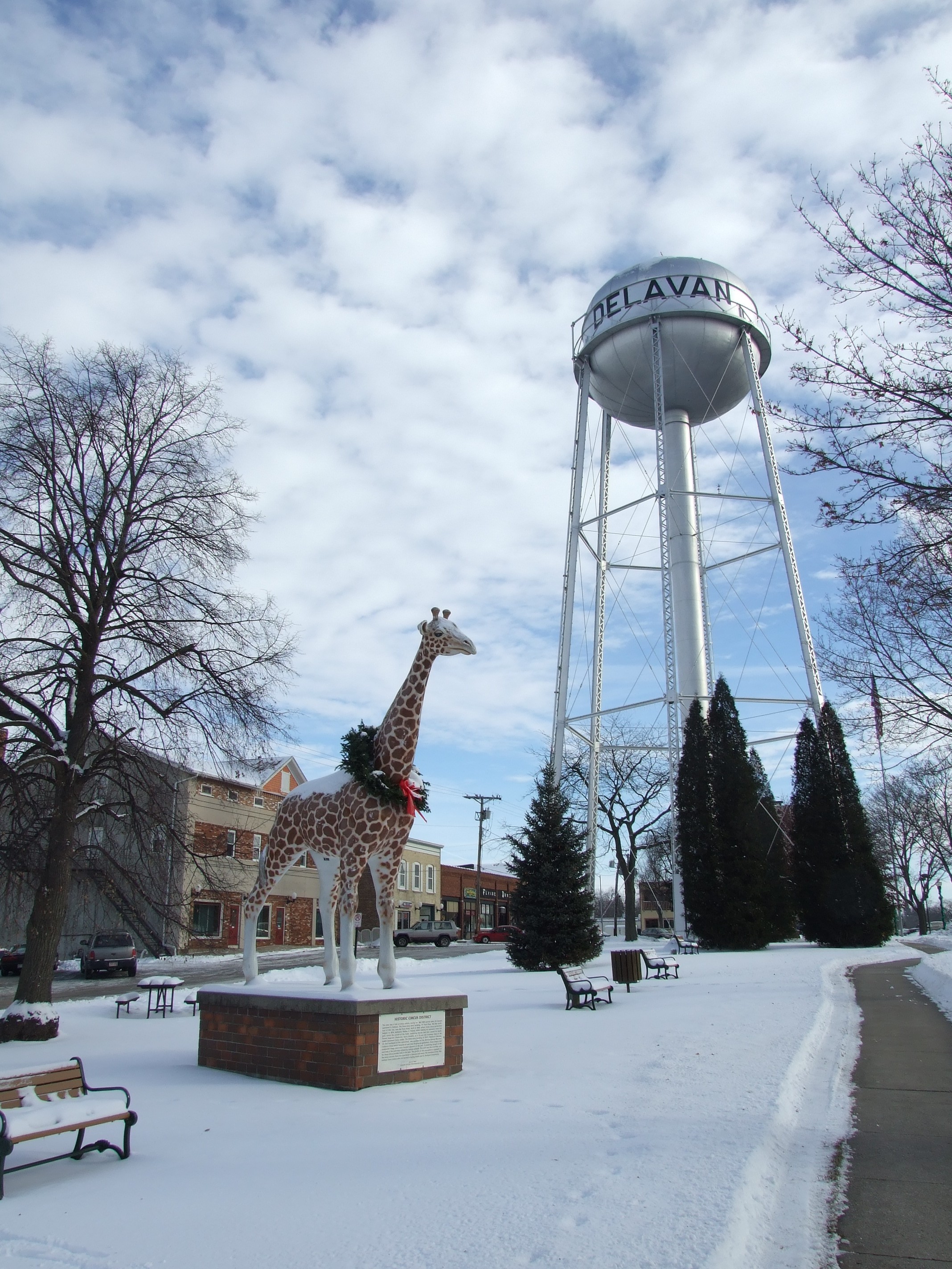 A giraffe statue on a brick pedestal beside a water tower and benches in a snow-covered park.
